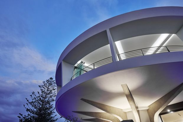 Low-angle exterior view of a sculptural multi-storey building with sweeping curved slab edges, recessed balconies, dark metal railings, and dramatic branching structural columns beneath. The facade is predominantly pale rendered concrete or plaster, set against a blue evening sky.