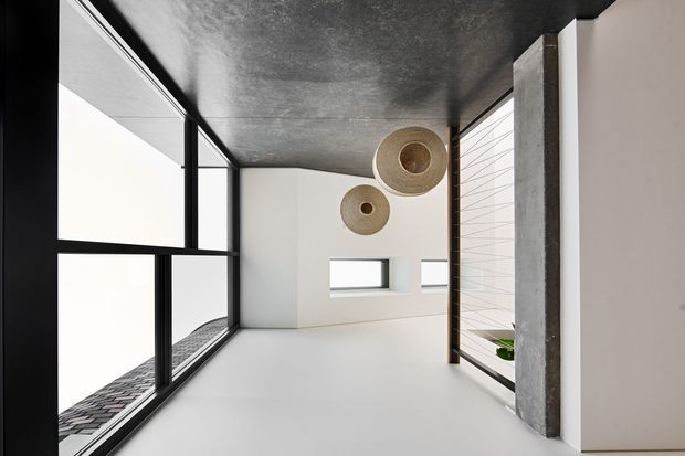 A minimalist modern interior passage or living space with extensive black-framed glazing, white walls, a dark textured ceiling, and a prominent exposed concrete column. Two woven pendant lights hang above the open area, with a glimpse of patterned exterior paving and greenery beyond the glazing.