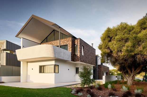 Contemporary multi-level residential exterior with a projecting upper floor, broad cantilevered balcony, exposed brick feature walls, smooth white rendered walls, dark metal cladding, and a sharply pitched roof. The home is set within a landscaped suburban garden with mature trees and low planting.
