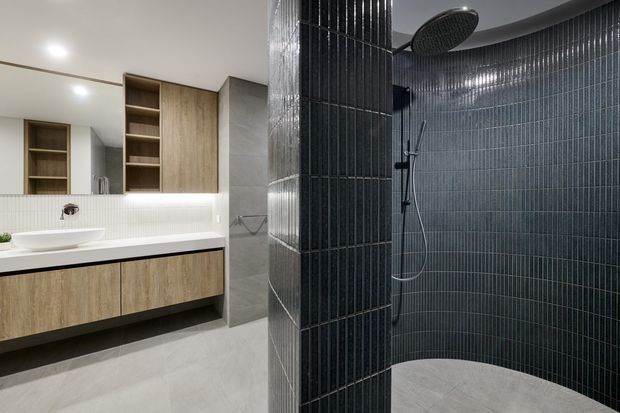 Contemporary bathroom featuring a floating timber-look vanity with white basin and benchtop, open wall shelving, large grey floor and wall tiles, and a curved shower enclosure lined with narrow dark vertical tiles. Matte black shower fixtures and a large round overhead shower add contrast.