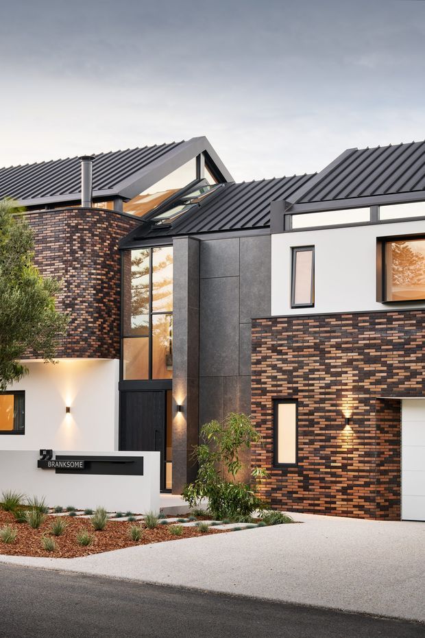 A contemporary multi-level residential facade viewed from the street, featuring dark standing-seam metal gabled roofs, richly patterned brown and charcoal brickwork, white rendered walls, charcoal cladding panels, large glazed openings, and a landscaped entry with a Branksome address sign. The low-maintenance planting and illuminated facade create a refined modern street presence.