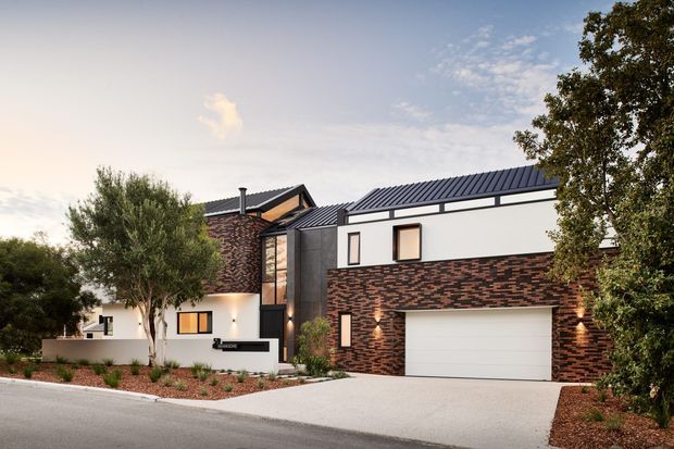 Contemporary multi-level residential house viewed from the street, featuring dark standing-seam pitched roofs, white rendered walls, expressive red-brown patterned brickwork, black-framed windows, a glazed stairwell or entry volume, and an integrated double garage. The landscaped front garden includes mature trees, low planting, and a broad pale driveway.