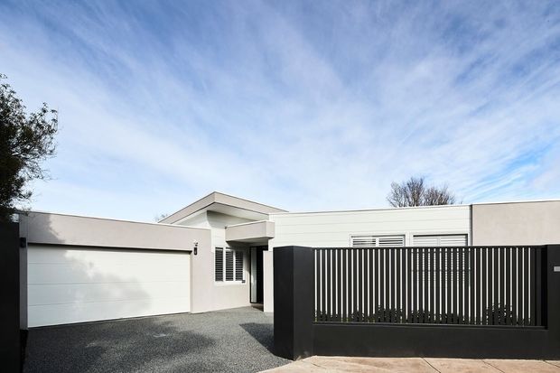Street-facing view of a low, single-storey contemporary residence with a flat roof, white rendered walls, an integrated double garage, and a prominent black vertical-slat privacy fence and gate. The driveway is finished in dark aggregate, with a small covered entry visible behind the fence.