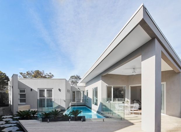 Exterior architectural photograph of a contemporary single-storey residence with a sharply angled roofline, white rendered walls, extensive glazing, and a covered outdoor living area. A swimming pool and timber-toned deck sit within the courtyard, with sculptural plantings along the edge.