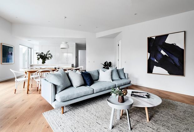 Bright open-plan contemporary living and dining area with white walls, pale timber flooring, a large light-grey sectional sofa, layered round coffee tables, a textured neutral rug, and abstract wall artwork. A white kitchen with island seating and pendant lighting is visible beyond the dining area.