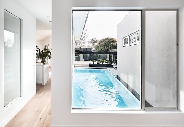 Bright contemporary interior view looking through a large glazed opening toward a narrow outdoor swimming pool and enclosed courtyard. The room has white walls, pale timber flooring, minimalist built-in cabinetry, and a large wall mirror or reflective panel. The pool, rendered exterior walls, dark fencing, and planting are visible beyond the glazing.