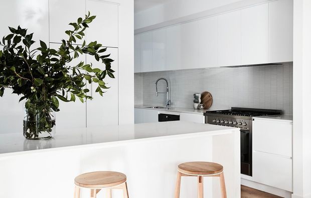 Bright contemporary white kitchen with a large white island, integrated white cabinetry, a fine grey mosaic tile splashback, stainless steel range cooker and sink, and light timber stools. A large leafy arrangement in a glass vase adds an organic accent.