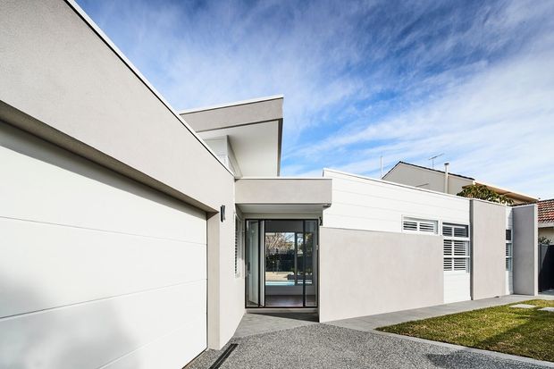 Contemporary low-rise residential facade with crisp white and light-grey rendered walls, a flat roofline, large glazed entry, horizontal louvred windows, and a broad white garage door. A gravel driveway and small lawn sit in front, with blue sky above.