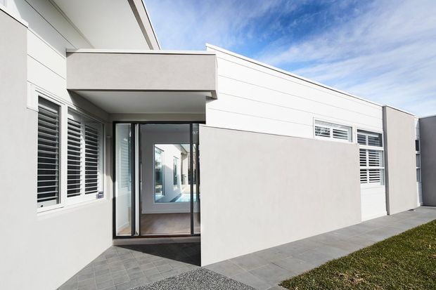 Contemporary single-storey residential facade with smooth light rendered walls, strong horizontal lines, white plantation shutters, a glazed entry, and a grey tiled pathway. The building presents a clean, minimalist exterior under a bright sky.