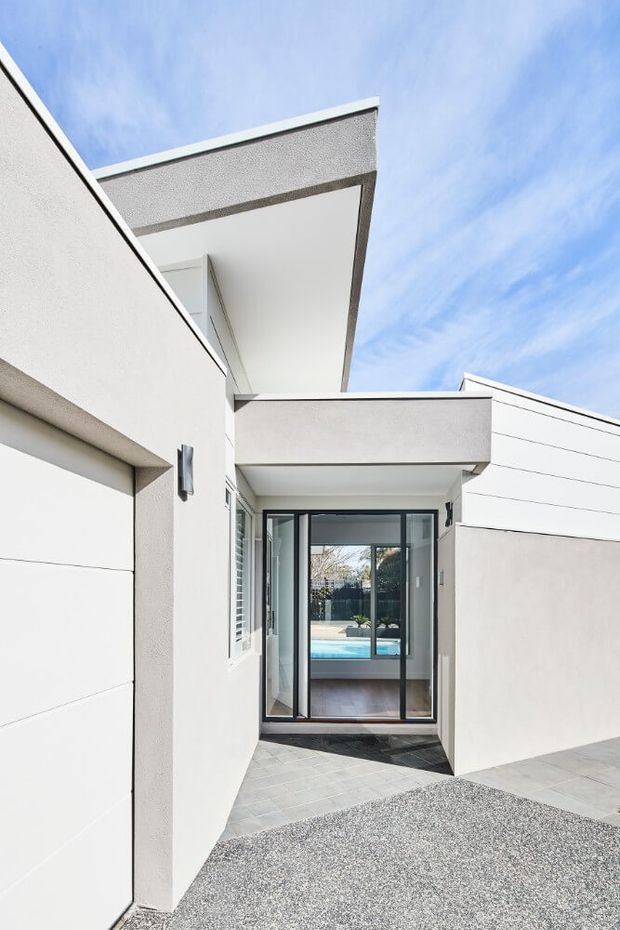 A bright contemporary residential exterior courtyard or entry passage with white rendered walls, crisp angular roof overhangs, horizontal cladding, dark-framed glazed doors, and pale tiled paving. Through the glazing, a timber-floored interior and blue swimming pool are visible. The composition emphasizes clean geometric forms and a sheltered connection between the driveway and the home.