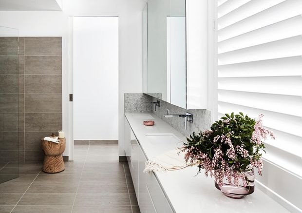 Bright contemporary bathroom with a long white vanity, integrated basin, mirrored wall cabinets, grey stone-look splashback, large grey floor and wall tiles, and a wide white plantation-shutter window. A freestanding toilet is partially visible at the rear, with decorative flowers on the vanity.