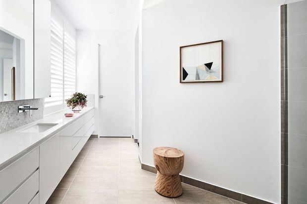 Bright, minimalist bathroom with a long white floating vanity, integrated double basins, pale mosaic splashback, large horizontal mirror, plantation shutters, and pale tiled flooring. A frameless shower enclosure is visible at right, with simple geometric artwork and a sculptural timber stool adding subtle detail.