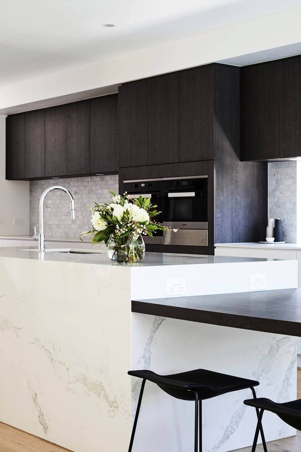 Contemporary kitchen with dark timber cabinetry, integrated black appliances, a pale stone island and splashback, and a black breakfast-bar extension with stools. A chrome gooseneck tap and flower arrangement sit on the island, while white walls and recessed lighting keep the space bright.