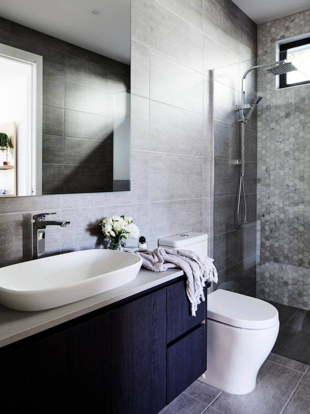 Contemporary bathroom featuring a dark timber-look floating vanity with a pale stone benchtop, white vessel basin, wall-mounted mirror, large grey wall and floor tiles, and a glass-enclosed shower with a rain showerhead. A patterned mosaic tile feature wall surrounds the black-framed window, while a white toilet and soft textile accents complete the space.