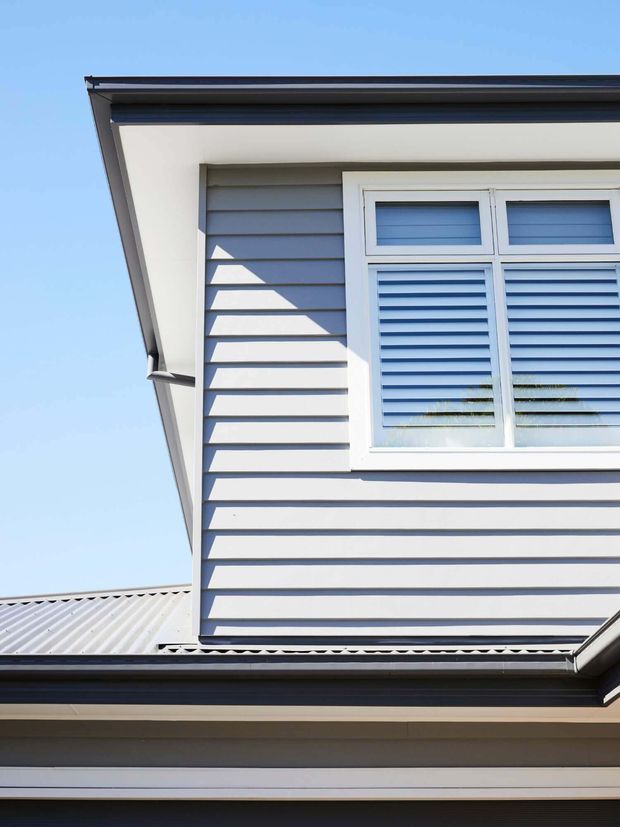 Close-up exterior view of a two-storey residential facade with blue-grey horizontal weatherboard cladding, white trim, a louvred window, dark gutters and a corrugated metal roof. Strong sunlight creates crisp shadows across the cladding against a clear blue sky.