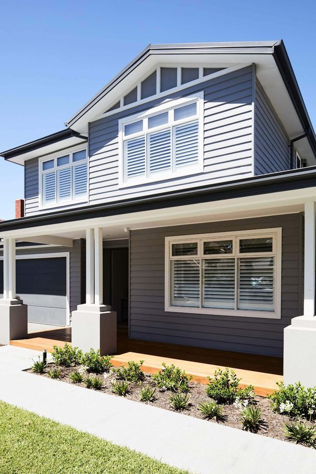 Exterior view of a two-storey suburban home with dark grey horizontal weatherboard cladding, white-trimmed windows with plantation shutters, a gabled roof, covered front verandah, attached garage, timber decking, and a small landscaped garden.