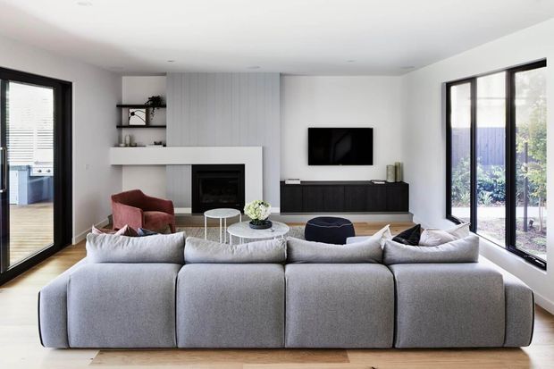 A bright, contemporary living room viewed from behind a large modular sectional sofa. The room features a white fireplace wall with vertical grey panelling, open shelving, a wall-mounted television, dark low-profile media cabinetry, black-framed glazing, and light timber flooring. A small glimpse of an adjacent outdoor area and another room is visible through the glazing.