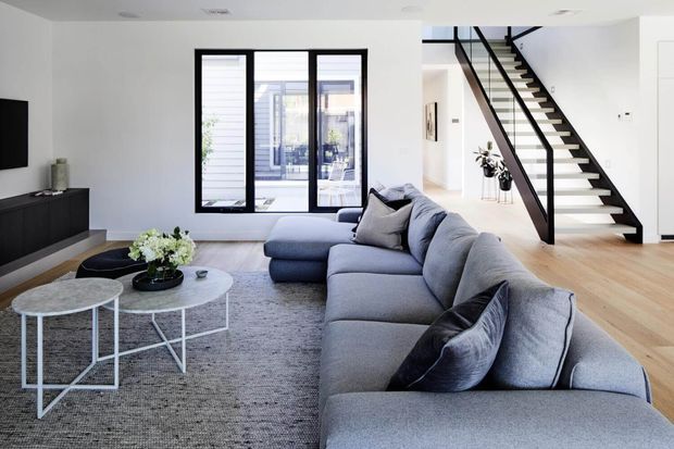 A bright, contemporary open-plan living room with a large L-shaped grey sectional sofa, nested round coffee tables, a textured grey rug, and minimalist black media cabinetry. Black-framed glazing opens the room visually to an outdoor area, while a striking open-riser staircase with black steel rails leads upward along the rear wall. Pale timber flooring and white walls create a clean, airy interior.
