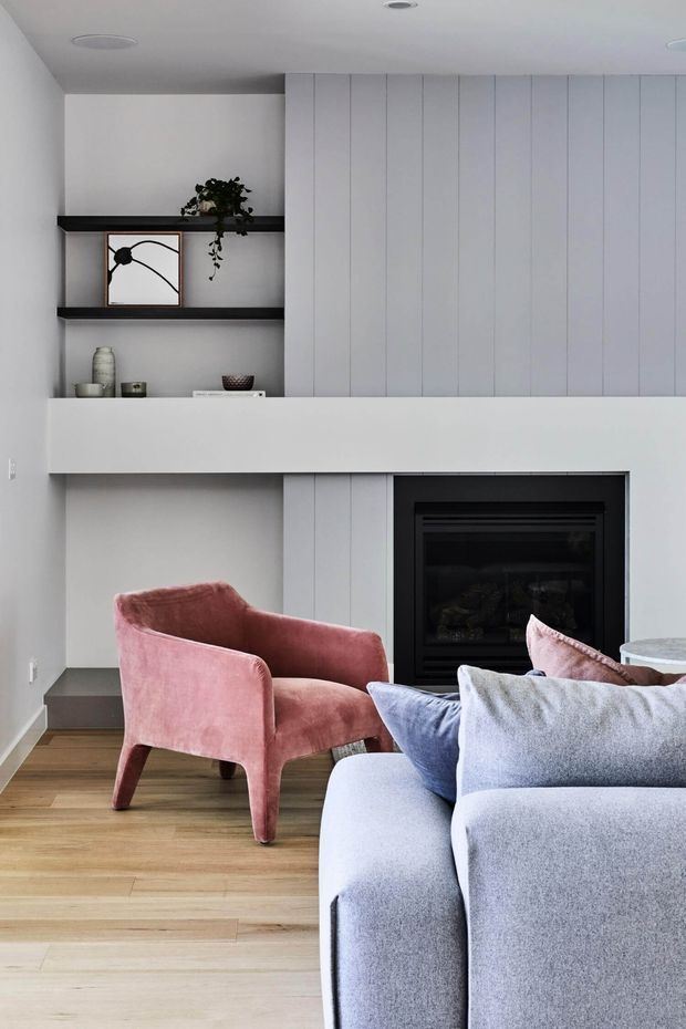 A contemporary living room featuring a built-in black fireplace, white vertical-panel cabinetry and shelving, pale timber flooring, a blush pink lounge chair, and a large pale grey sectional sofa with blue and pink cushions. The room has a restrained, modern palette with soft decorative accents.