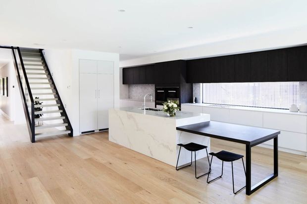 Bright open-plan contemporary kitchen and dining area with pale timber flooring, a large white stone island, dark upper cabinetry, white lower cabinetry, integrated appliances, and a black dining table with stools. An open-riser staircase with black framing leads upward beside the kitchen.