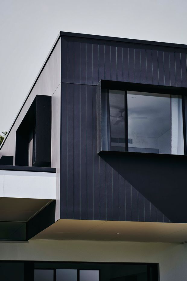 Close-up architectural photograph of a contemporary multi-level building facade. The design features dark vertically ribbed cladding, crisp geometric projecting forms, black-framed glazing, white rendered walls, and a flat or very low-pitched roofline. The composition emphasizes strong cantilevered and angular volumes with a restrained monochromatic palette.