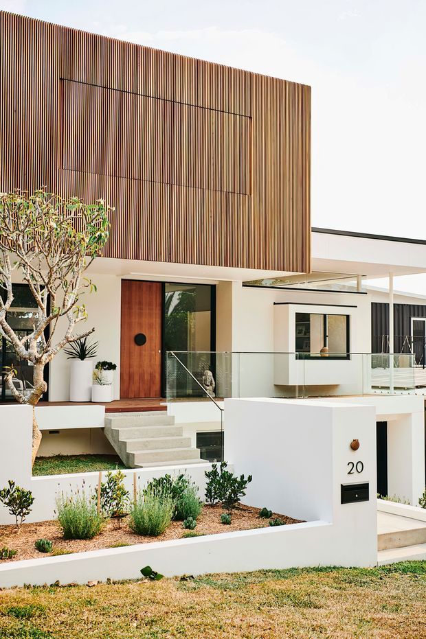 Street-facing view of a contemporary multi-level residence with a strong geometric facade. The upper storey is clad in closely spaced vertical timber battens, contrasting with smooth white rendered walls and dark-framed glazing. A timber pivot entry door opens onto a raised landing with frameless glass balustrades and concrete steps. The landscaped front garden uses low, drought-tolerant planting and gravel, while a white rendered entry wall displays the number 20 and an integrated letterbox.
