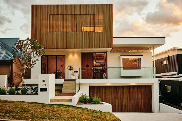 A contemporary two-storey residential facade on a sloping site, featuring a projecting upper volume clad in vertical timber battens, white rendered walls, extensive glazing, a sheltered entry, and a large timber-toned garage door. The home sits among neighbouring houses under a warm evening sky.