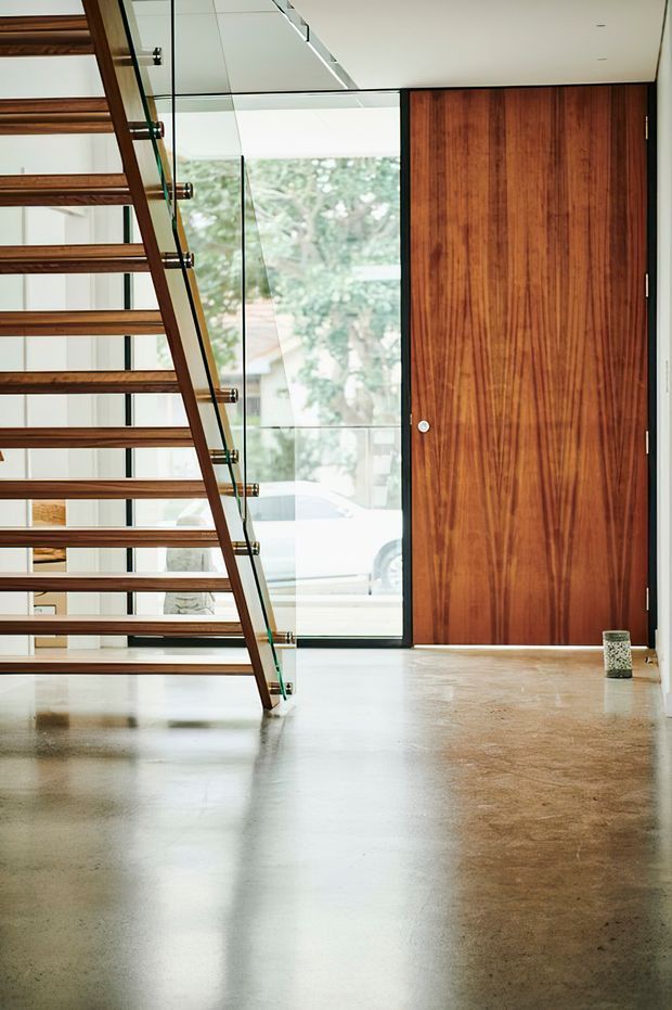 A bright modern interior entry space featuring a floating timber staircase with glass balustrades, a large richly grained timber pivot door, full-height glazing, white walls and ceiling, and a polished concrete floor. The composition emphasizes clean lines, natural materials, and abundant daylight.
