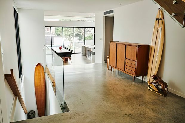 A bright, open-plan modern interior viewed from an upper-level landing or hallway. The space features polished concrete flooring, white walls, black-framed glazing, a glass stair balustrade, and a mid-century wooden sideboard. Surfboards and wooden boards are displayed along the wall and stair opening, while a dining area and living space are visible beyond.