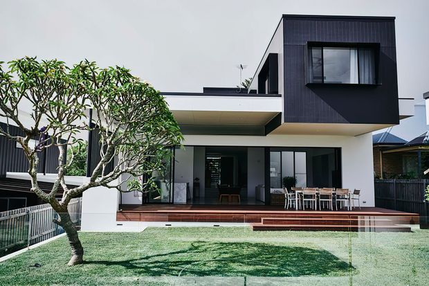 A contemporary two-storey residential house viewed from the landscaped front yard, with a crisp white rendered ground floor and a projecting upper volume clad in dark vertical panels. Large sliding glass doors open onto a broad timber deck with outdoor dining furniture, while a mature frangipani tree and lawn occupy the foreground. Strong cantilevered forms, flat rooflines, and extensive glazing define the facade.