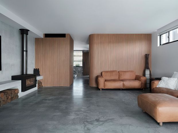 A contemporary open-plan living room with a polished grey concrete floor, white walls and ceiling, and extensive vertical timber slat feature walls forming partial room dividers and concealed joinery. A black wood-burning fireplace sits against the left wall, while tan leather sofas and an ottoman furnish the room. High horizontal windows bring in natural light, and a glimpse of an adjoining space is visible through the slatted partition.