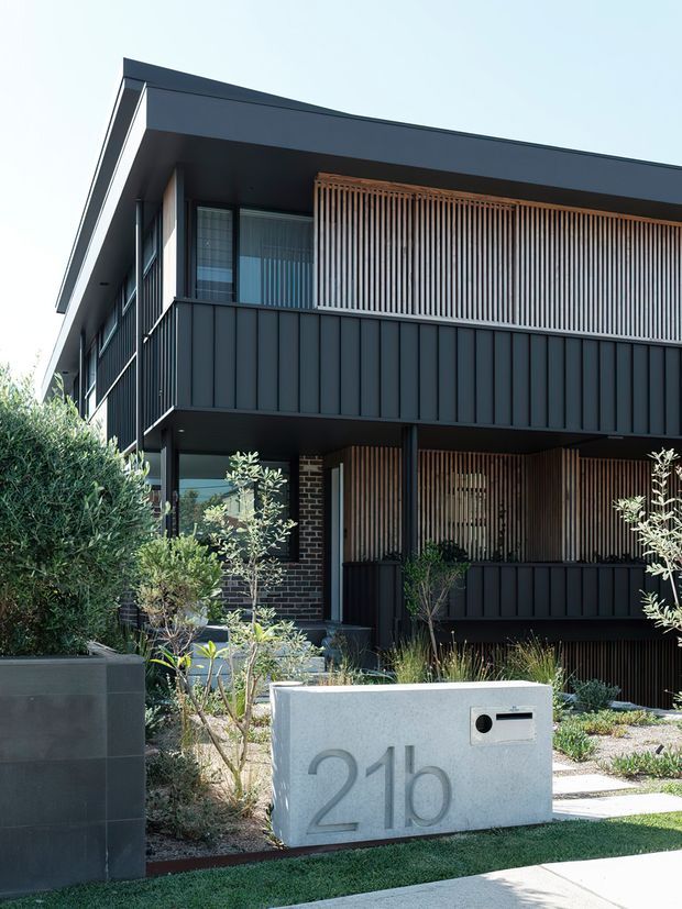 A contemporary multi-storey residential facade viewed from the front garden. The dark-clad building has a steep asymmetrical roof edge, projecting balconies, vertical metal wall panels, extensive glazing, and timber-toned vertical privacy screens. Native-style landscaping and a large concrete address monument sit in the foreground.