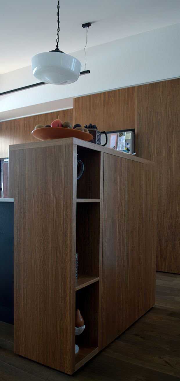 Interior view of a warm, wood-lined kitchen or open-plan living area, focused on a tall timber kitchen island or built-in storage unit with open shelving. Dark wood flooring and extensive walnut-toned cabinetry are paired with a white ceiling and a sculptural white pendant light. A long black linear light fixture and colourful decorative objects add contrast.