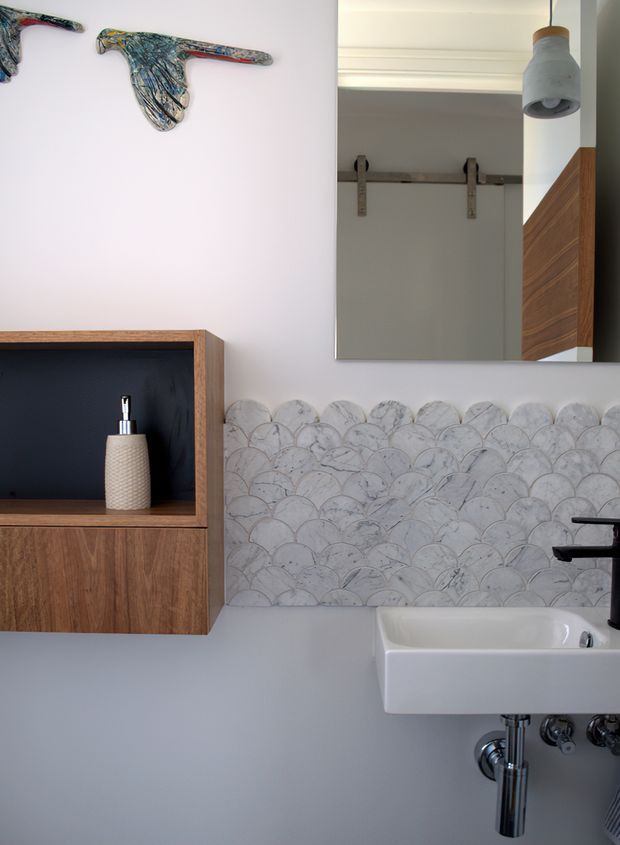 A refined contemporary bathroom featuring a wall-mounted white basin with exposed chrome plumbing, a timber wall cabinet with an open dark niche, and a decorative scalloped marble tile splashback. A large mirror reflects a barn-style sliding door and timber wall detail, while a pendant light and sculptural wall artwork add character.