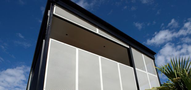 Low-angle exterior view of a modern upper-level balcony or verandah enclosed with large white mesh privacy screens and framed by dark structural elements. The building has a clean, angular form under a deep blue sky, with palm foliage at the edge of the frame.