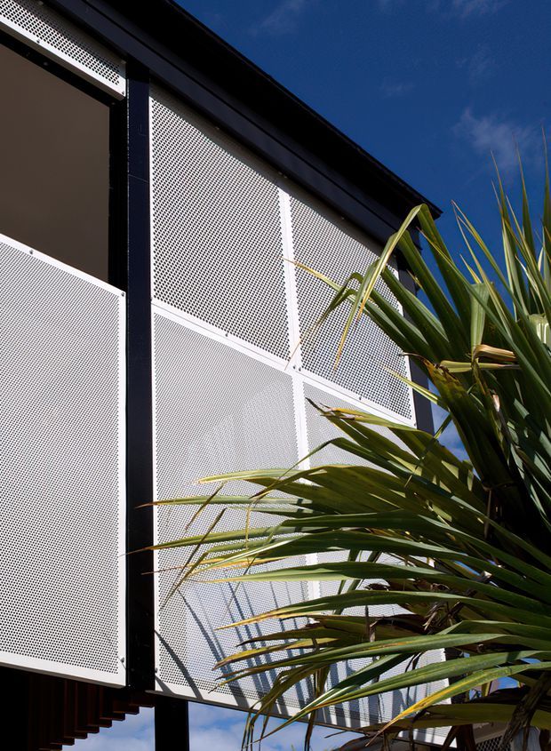 Upward-looking exterior detail of a contemporary building facade featuring white perforated metal screening panels framed by dark structural elements. Strong sunlight, deep blue sky, and dense spiky green planting create a high-contrast architectural composition.