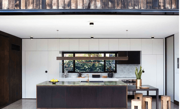 Contemporary kitchen interior with a large dark island, white flat-panel cabinetry, pale tiled splashback, timber wall panelling, black-framed horizontal window, and a long suspended timber pendant. A dining/work table and stools sit behind the island, with greenery visible through the window.