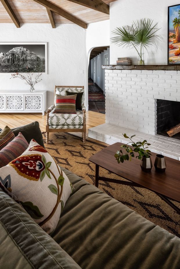 Warm, eclectic living room with a white-painted brick fireplace, exposed timber ceiling beams, white textured walls, and honey-toned timber flooring. A deep olive sofa with embroidered cushions faces a dark timber coffee table, while a patterned armchair, woven rug, carved white console, artwork, and plants add layered character. An arched hallway opening extends from the room.