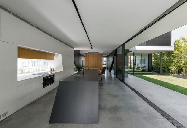 A sleek, minimalist open-plan kitchen viewed from the island, with white built-in cabinetry, a dark island benchtop, integrated appliances, warm timber wall panelling, polished concrete flooring, and full-height sliding glass doors opening to a landscaped courtyard. The space has a long, low contemporary form with strong indoor-outdoor connection and abundant natural light.