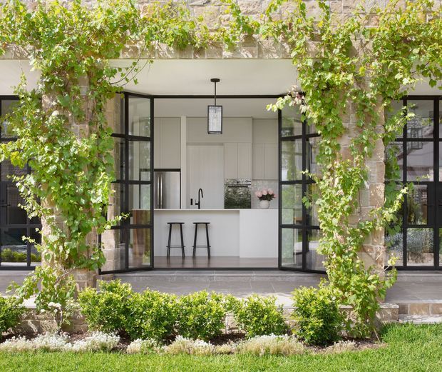 A bright, refined kitchen viewed from a landscaped courtyard through open black-framed glazed doors. The compact white kitchen has a slim island with two stools, minimalist cabinetry, a dark benchtop, and a pendant light. Stone masonry, climbing greenery, manicured hedges, and a paved terrace frame the indoor-outdoor composition.