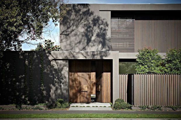 Street-facing contemporary two-storey residence with a minimalist rectilinear facade. The home combines smooth light-grey rendered walls, dark recessed openings, vertical timber or timber-look screening, a large timber pivot entry door, and a tall slatted privacy fence. Carefully clipped planting and mature trees soften the strong geometric architecture.