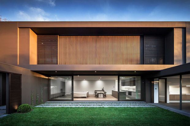 Contemporary two-storey residence with a dark, rectilinear facade and deep recessed upper-level openings. Warm vertical timber screening forms a central feature, while extensive ground-floor glazing opens toward a landscaped courtyard with layered stone paving and lawn. The composition is photographed externally at dusk, with warm interior lighting visible through the glass.