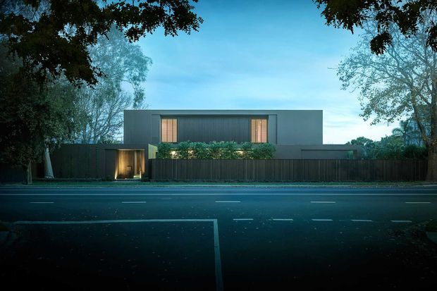 A dusk street view of a minimalist, single-storey modern residence behind a tall dark vertical-slat fence. The flat-roofed, rectilinear facade has dark grey cladding or rendered walls, narrow warm-lit timber-toned windows, a landscaped hedge, and a recessed illuminated entry gate. Mature trees frame the property, with a roadway in the foreground.