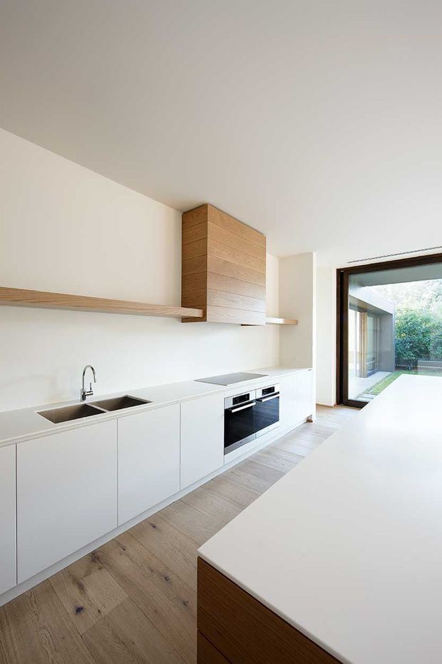 A bright, minimalist contemporary kitchen with white handleless cabinetry, integrated black appliances, a pale engineered-stone benchtop, stainless-steel sink and tap, and light timber flooring. A large timber-clad overhead joinery volume and floating timber shelf add warmth. Full-height glazing opens toward a landscaped courtyard, while a large island occupies the foreground.