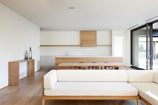 Bright open-plan interior combining a minimalist kitchen, dining area and living room. The space features white walls and cabinetry, light timber flooring and joinery, a long kitchen island with timber detailing, a built-in dining bench, and a large white sectional sofa. Full-height black-framed glazing opens to the right, bringing in abundant natural light.