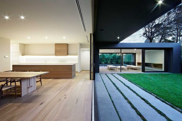 A contemporary open-plan kitchen and dining area opens directly onto a landscaped courtyard through full-height glazing. The interior features pale timber flooring, white cabinetry, a timber kitchen island and dining table, while the exterior has a dark flat-roofed pavilion, concrete stepping slabs, lawn, and a sheltered outdoor seating area.