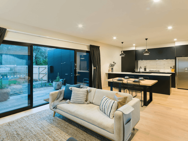 Open-plan living and kitchen area with a light upholstered sofa, textured rug, timber flooring, and a compact dining table. The room features large black-framed sliding glass doors opening to an outdoor area, matte black kitchen cabinetry, a white tiled splashback, stainless-steel appliances, pendant lighting, and warm recessed ceiling lights.