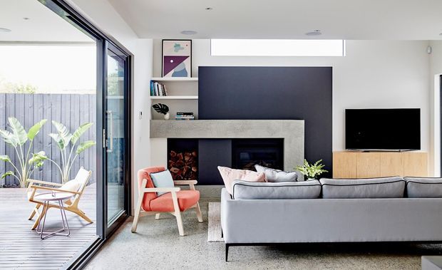 Bright contemporary living room with a large grey sectional sofa facing a wall-mounted television and timber media console. A textured stone fireplace surround with dark feature backing anchors the room, while full-height sliding glass doors open to a timber deck and planted outdoor area. The space has white walls, recessed lighting, and colourful modern artwork and accent chairs.