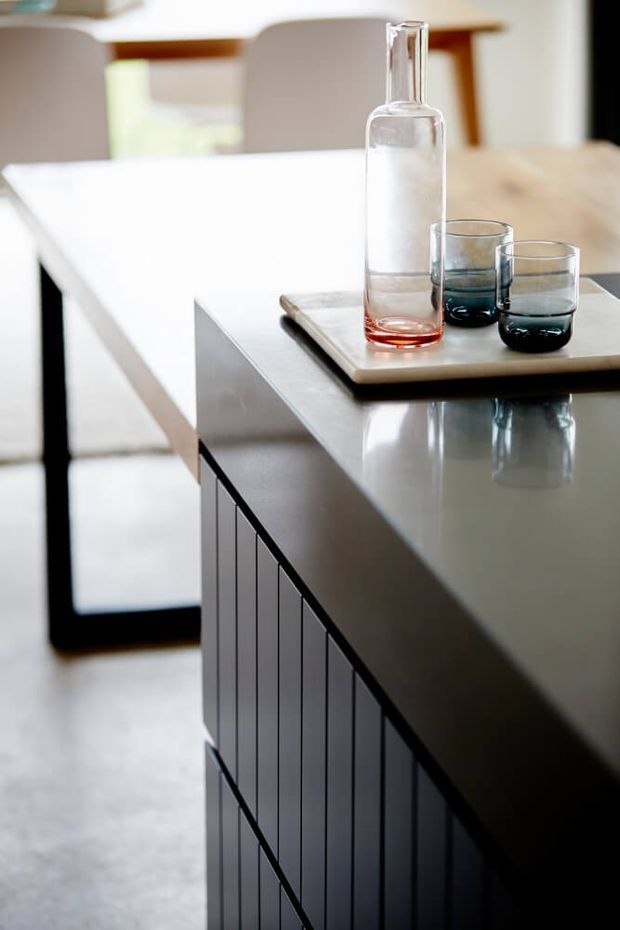 Close-up interior view of a contemporary kitchen island with a dark, vertically fluted or panelled base and a smooth light-grey benchtop. A wooden serving board with a clear glass bottle and tumblers sits on the island. A dining table and upholstered chairs are softly visible in the background, with bright natural light and reflective surfaces creating a clean, refined atmosphere.