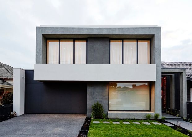 Front exterior view of a contemporary two-storey residence with a flat roof, strong geometric massing, exposed grey concrete-look facade panels, dark garage and entry elements, large upper-level windows with a broad white balcony parapet, and a landscaped lawn with stepping stones.